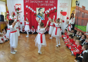Dziewczynki tańczą po kole z uniesionymi do góry rękoma. W dłoniach trzymają biało-czerwone kwiaty.