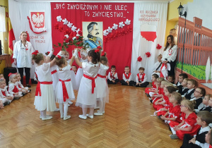 Dziewczynki tańczą w kółeczku z uniesionymi do góry rękoma. W dłoniach trzymają biało-czerwone kwiaty.