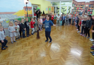 Dzieci tańczą kole. W srodku koła chłopiec tańczy z miotłą.