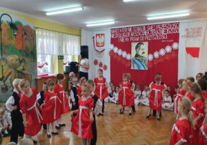 Dziewczynki w czerwonych sukienkach, oraz chłopcy w strojach galowych tańczą Mazura.