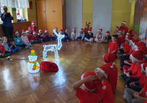 Dzieci siedzą na sali. Na środku znajdują się świecący renifer i bałwan.