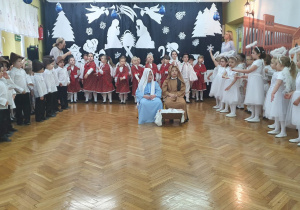 Na środku sceny siedzą dzieci odgrywające rolę Józefa, Maryji. Wokół nich wszystkie przedszkolaki śpiewają pastorałkę.