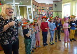 Dzieci z "owocowymi kapeluszami" na głowach wraz z prowadzącą grają na instrumentach.