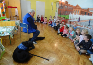 Dzieci siedzą, naprzeciw nich pies z terapeutą.