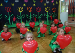 Dzieci kucają w rzędach, w rękach trzymają czerwone balony w kształcie serc.