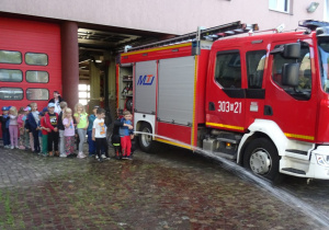 Dzieci stoją w kolejce przy wozie strażackim. Z pomocą strażaka polewają wodą kostkę.