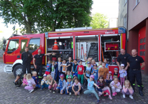 Dzieci wraz ze strażakami pozują do zdjęcia przy wozie strażackim.