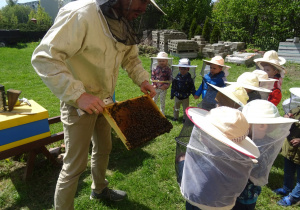 Dzieci w strojach pszczelarskich, pszcelarz i ul. Pszczelarz prezentuje ramkę z pszczołami