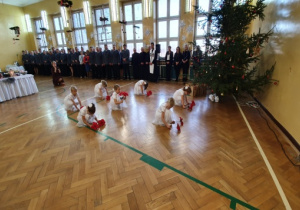 Dziewczynki w tańcu kucają z kwiatami - gwiazdami betlejemskimi, w tle służby mundurowe.