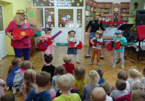 Czworo dzieci razem z prowadzącą koncert na środku sali gra na gitarach.