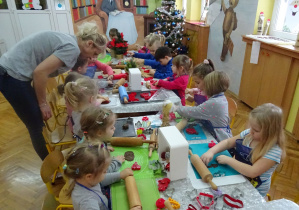 Dzieci przy ozdobionym świątecznie stole z pomocą nauczycielki rozwałkowują ciasto na pierniki.