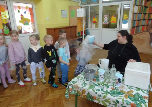Dzieci stojące w kolejce, dziewczynka siedzi na krzesełku podczas doswiadczenia z suchym lodem.