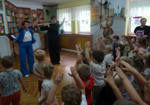 Prowadząca i przedszkolaki trzymają ręce uniesione do góry.