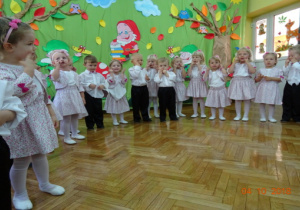 Ubrane odświętnie dzieci śpiewają piosenkę trzymając się za policzki.