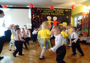 Na scenie na tle dekoracji z okazji Dnia Babci i Dziadka tańczą dzieci w parach oraz nauczycielka.