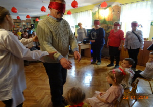 Wnuczęta siedzą na krzesełkach. 1dziadek z zasłoniętymi oczyma próbuje wskazać swojego wnuczka. Obok stoja babcie z zasłoniętymi oczyma, które czekają na swoją kolej.