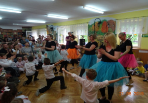 Mamusie w czarnych podkoszulkach i kolorowych spódnicach wykonują taniec wspólnie ze swoimi synami.