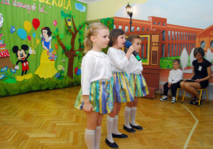 Na tle dekoracji z tortem i bajkowymi postaciami stoją trzy dziewczynki. Z boku siedzi nauczycielka z chłopcem.