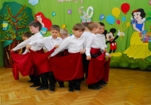 Na tle dekoracji z tortem i bajkowymi postaciami w kółeczkiu są chłopcy z bordowymi chustami. Dzieci ykonują układ taneczny Flamenco.