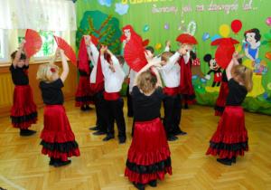 Na tle dekoracji z tortem i bajkowymi postaciami chłopcy i dziewczęta z wachlarzami wykonują taniec Flamenco. W środku stoją chłopcy z chustami w górze oraz dziewczęta trzymające w górze wachlarze.
