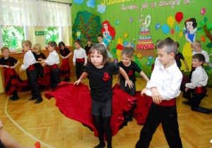 W dewóch rzędach stoją dzieci tańczącr flamenco. Na pierwszym planie dziewczynka zrzucająca spódnicę.
