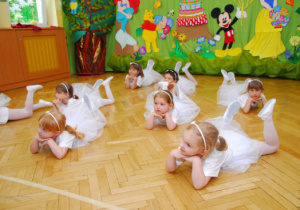 Na tle dekoracji z tortem i bajkowymi postaciami dziewczynki w białych sukienkach i białych opaskach leżą na brzuchu, ręce mają podparte pod brodą.
