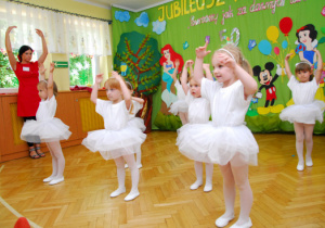 Na tle dekoracji z tortem i bajkowymi postaciami dziewczynki w białych sukienkach tańczą z ekoma uniesionyumi do góry.