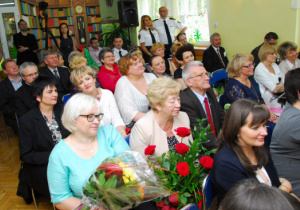 Na krzesełakach siedzą zgromadzeni goście, a niektórzy stoją. jedna z kobiet trzyma kwiaty.