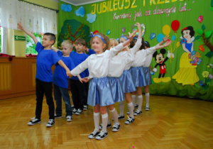 Na tle dekoracji z tortem i bajkowymi postaciami dzieci tańczą w parch z rękoma uniesionymi do góry.